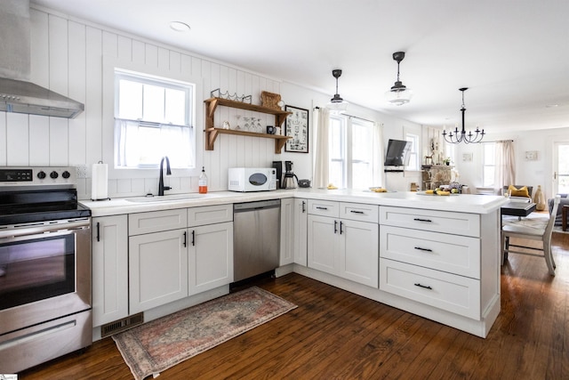 kitchen featuring a notable chandelier, white cabinets, wall chimney exhaust hood, appliances with stainless steel finishes, and decorative light fixtures