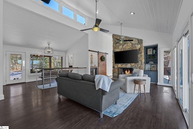 living room with dark hardwood / wood-style flooring, ceiling fan with notable chandelier, a barn door, high vaulted ceiling, and a stone fireplace