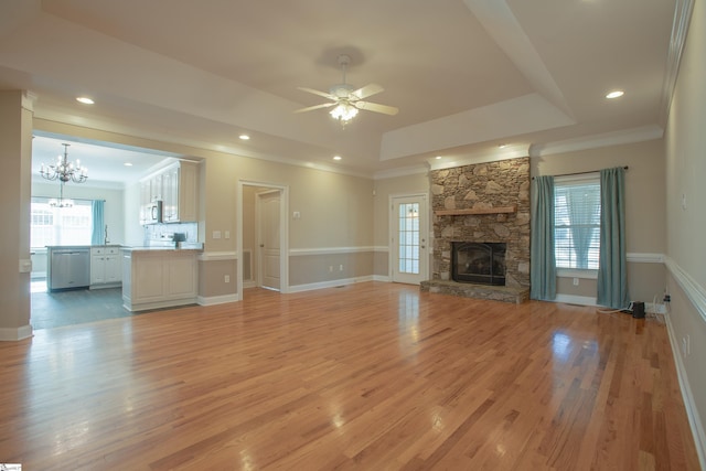 unfurnished living room featuring ceiling fan with notable chandelier, a stone fireplace, crown molding, light hardwood / wood-style flooring, and a tray ceiling