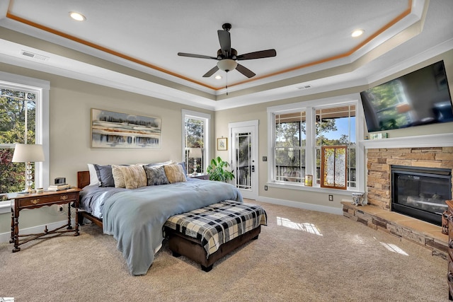 bedroom featuring light colored carpet, a raised ceiling, ceiling fan, crown molding, and a stone fireplace