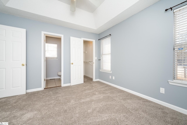 unfurnished bedroom featuring a walk in closet, ensuite bath, light colored carpet, a tray ceiling, and ceiling fan