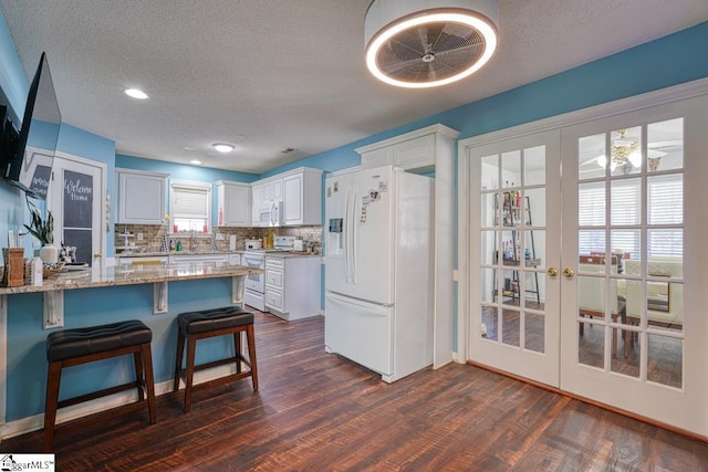 kitchen featuring light stone countertops, backsplash, white appliances, a kitchen bar, and white cabinets