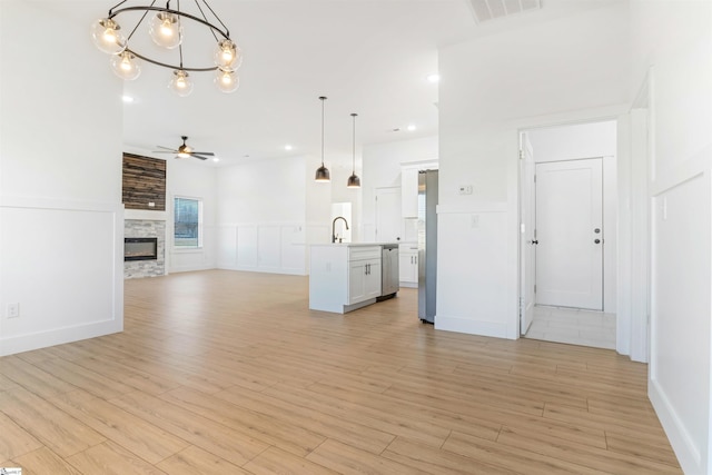 unfurnished living room featuring a stone fireplace, sink, ceiling fan with notable chandelier, and light wood-type flooring