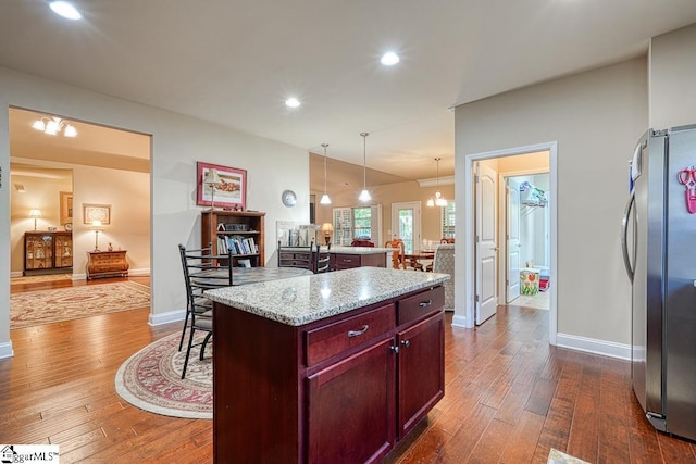 kitchen with decorative light fixtures, light stone counters, stainless steel refrigerator, a kitchen island, and dark wood-type flooring