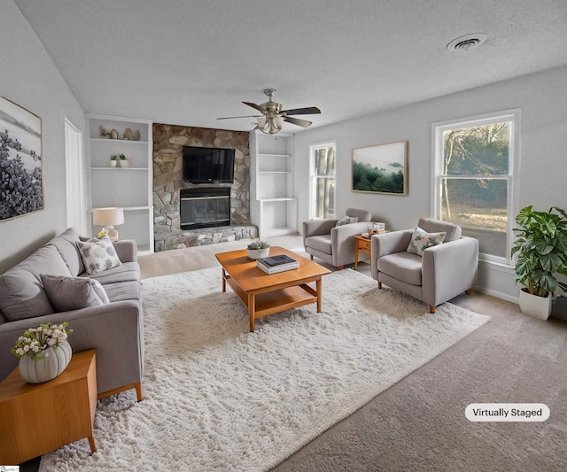 carpeted living room with a textured ceiling, ceiling fan, a stone fireplace, and built in shelves