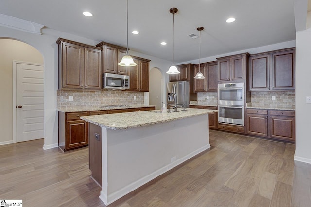 kitchen featuring stainless steel appliances, sink, decorative light fixtures, light stone countertops, and a kitchen island with sink