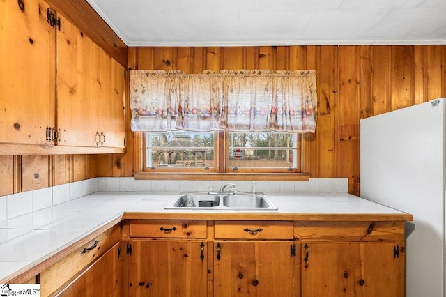 kitchen with tile countertops, white fridge, wood walls, ornamental molding, and sink