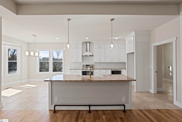 kitchen with white cabinets, light stone counters, an island with sink, stainless steel range, and wall chimney range hood