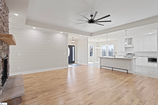 unfurnished living room featuring light wood-type flooring, a fireplace, a tray ceiling, sink, and ceiling fan with notable chandelier