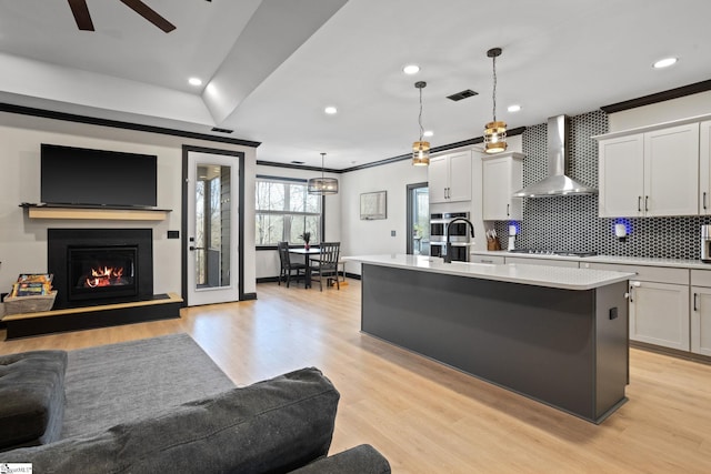 kitchen with pendant lighting, white cabinets, wall chimney range hood, light hardwood / wood-style floors, and a kitchen island with sink