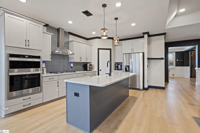 kitchen featuring appliances with stainless steel finishes, an island with sink, white cabinets, and wall chimney range hood