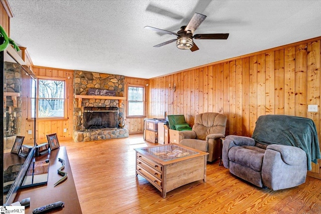 living area featuring a textured ceiling, a stone fireplace, light wood-style flooring, wood walls, and a ceiling fan