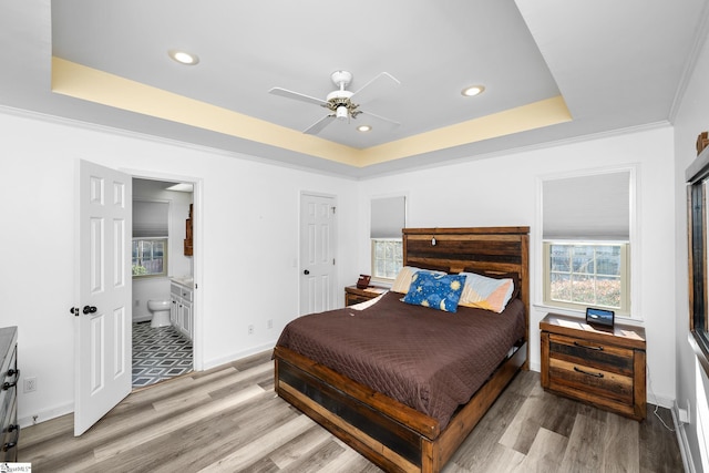 bedroom with ensuite bathroom, ceiling fan, a raised ceiling, light wood-type flooring, and multiple windows