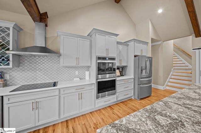 kitchen featuring tasteful backsplash, beamed ceiling, light wood-type flooring, stainless steel appliances, and wall chimney exhaust hood