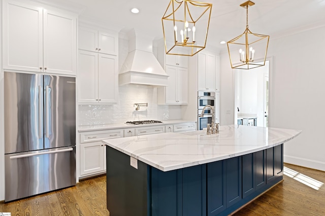 kitchen with white cabinets, appliances with stainless steel finishes, premium range hood, hanging light fixtures, and light stone counters