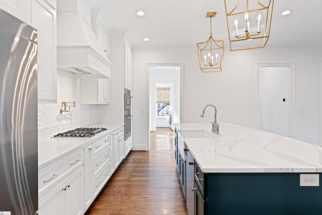 kitchen featuring white cabinetry, custom exhaust hood, appliances with stainless steel finishes, a kitchen island with sink, and hanging light fixtures