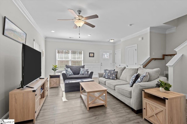 living area with wainscoting, ceiling fan, stairs, crown molding, and light wood-type flooring