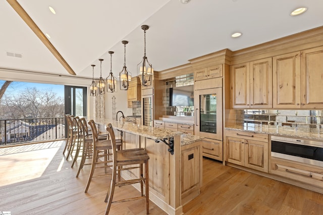 kitchen with lofted ceiling, decorative light fixtures, a center island with sink, light wood-type flooring, and light stone countertops