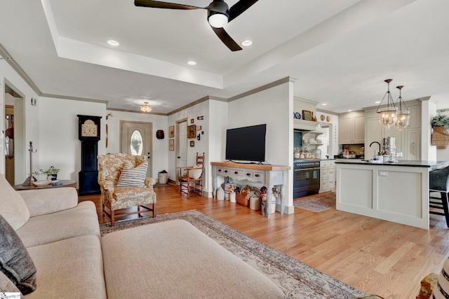 living room featuring sink, a tray ceiling, ceiling fan with notable chandelier, and light wood-type flooring