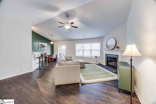 living area featuring lofted ceiling, dark wood-type flooring, a glass covered fireplace, a ceiling fan, and baseboards