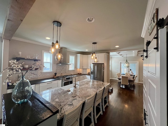 kitchen featuring wall chimney range hood, white cabinetry, backsplash, stainless steel appliances, and decorative light fixtures