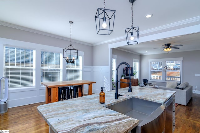 kitchen with pendant lighting, crown molding, light stone counters, a center island with sink, and dark hardwood / wood-style flooring