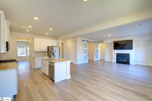 kitchen with appliances with stainless steel finishes, white cabinetry, light stone countertops, an island with sink, and a barn door