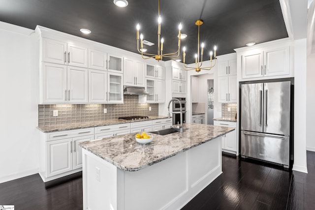 kitchen with light stone counters, appliances with stainless steel finishes, pendant lighting, a kitchen island with sink, and white cabinets