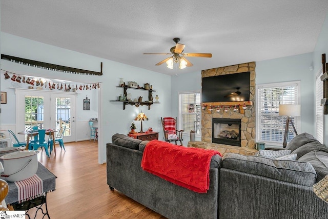 living room featuring light hardwood / wood-style flooring, ceiling fan, a textured ceiling, a stone fireplace, and french doors