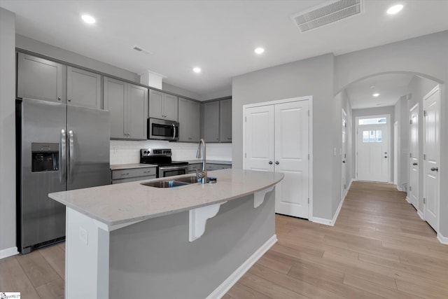 kitchen with sink, gray cabinetry, appliances with stainless steel finishes, an island with sink, and light stone countertops