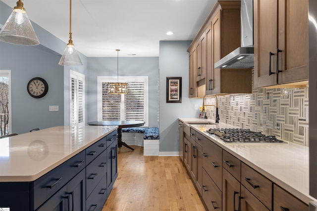 kitchen with wall chimney range hood, appliances with stainless steel finishes, hanging light fixtures, backsplash, and light hardwood / wood-style floors