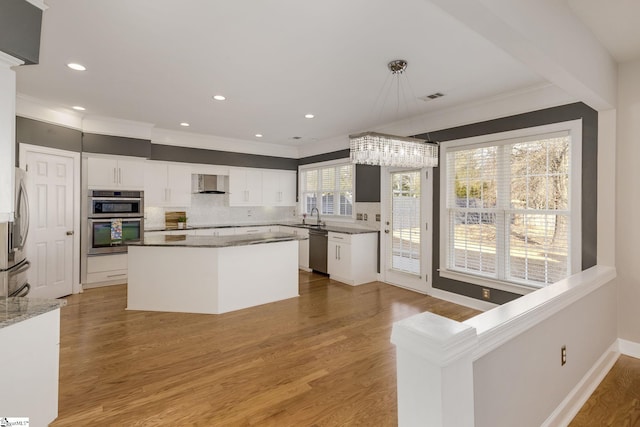 kitchen featuring wall chimney range hood, stainless steel appliances, hanging light fixtures, and white cabinets