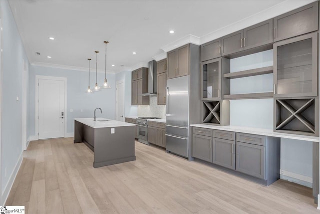 kitchen with wall chimney exhaust hood, hanging light fixtures, premium appliances, an island with sink, and light hardwood / wood-style floors