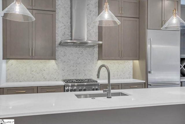 kitchen with wall chimney range hood, sink, built in refrigerator, hanging light fixtures, and light stone countertops