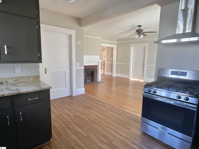 kitchen featuring wall chimney exhaust hood, stainless steel range with gas cooktop, ceiling fan, a fireplace, and light hardwood / wood-style floors