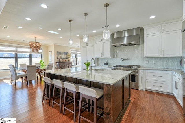 kitchen with wall chimney range hood, high end stainless steel range, white cabinets, and a large island with sink