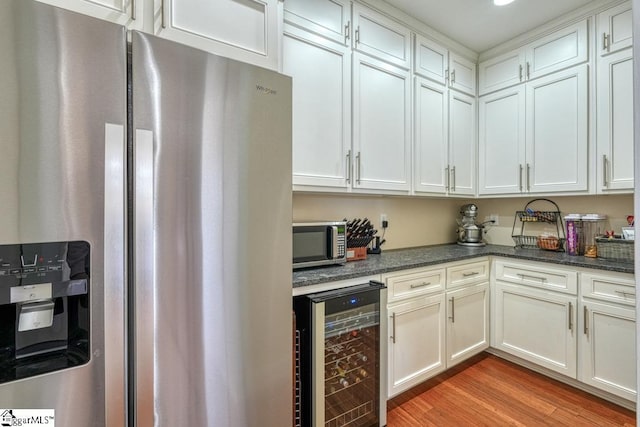 kitchen with appliances with stainless steel finishes, dark stone countertops, white cabinets, beverage cooler, and light hardwood / wood-style flooring