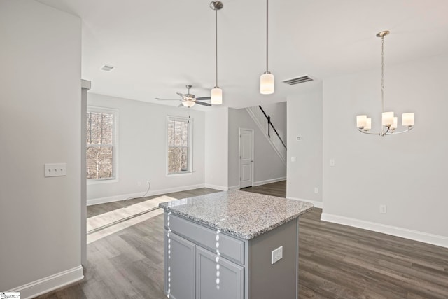 kitchen with dark wood-type flooring, a kitchen island, pendant lighting, light stone countertops, and ceiling fan with notable chandelier