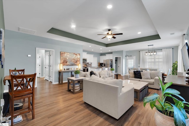 living room with a raised ceiling, ceiling fan with notable chandelier, and light hardwood / wood-style flooring