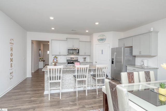 kitchen with hardwood / wood-style floors, stainless steel appliances, tasteful backsplash, light stone countertops, and a center island with sink