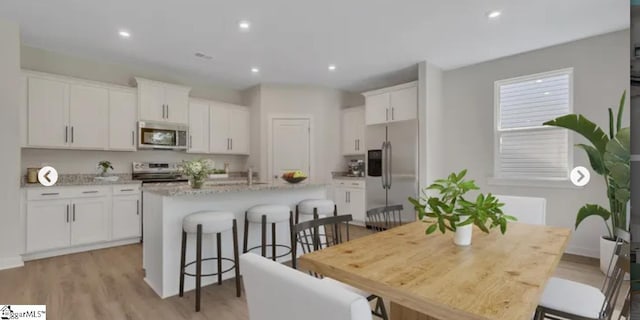 kitchen with appliances with stainless steel finishes, white cabinetry, an island with sink, a kitchen bar, and light stone counters