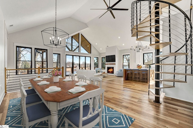 dining area featuring high vaulted ceiling, wood-type flooring, a fireplace, and ceiling fan with notable chandelier