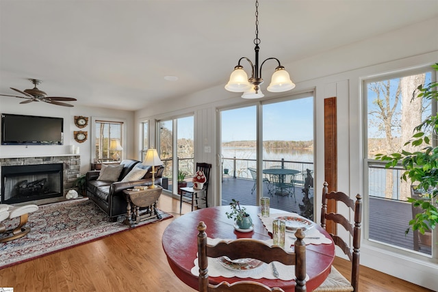 dining area featuring a stone fireplace, light wood-type flooring, ceiling fan with notable chandelier, and a water view