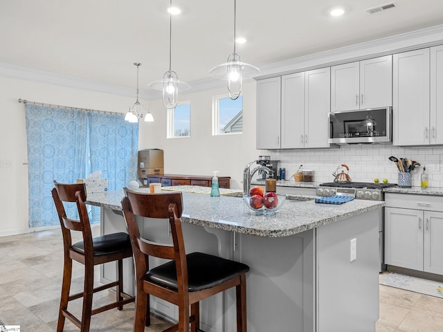 kitchen featuring hanging light fixtures, ornamental molding, stainless steel appliances, light stone countertops, and a kitchen island with sink