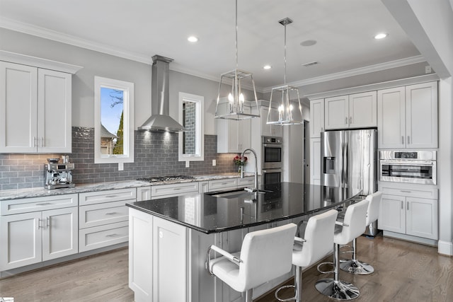 kitchen with wall chimney range hood, hanging light fixtures, white cabinets, and a center island with sink