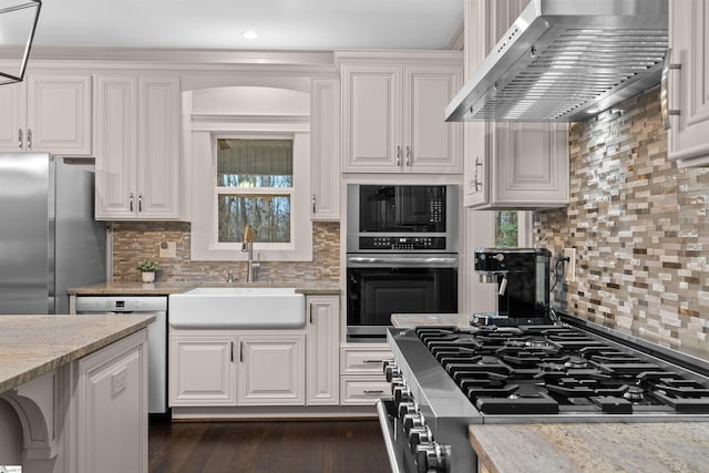 kitchen with sink, light stone counters, white cabinetry, stainless steel appliances, and wall chimney range hood