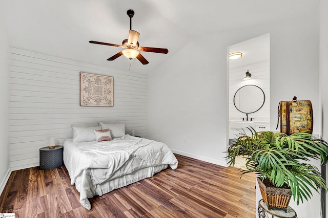 bedroom with ceiling fan, wooden walls, connected bathroom, wood-type flooring, and vaulted ceiling