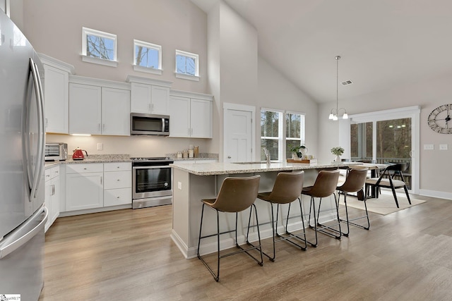 kitchen with light stone counters, decorative light fixtures, a center island with sink, appliances with stainless steel finishes, and white cabinets