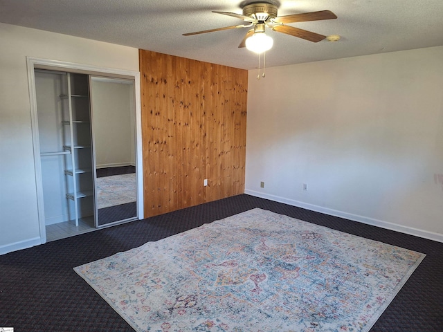 unfurnished bedroom featuring a ceiling fan, a textured ceiling, a closet, wooden walls, and dark colored carpet
