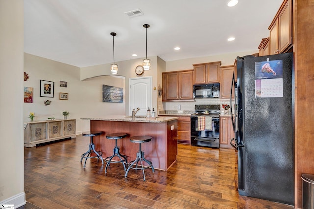 kitchen with a center island with sink, light stone counters, black appliances, sink, and a kitchen bar